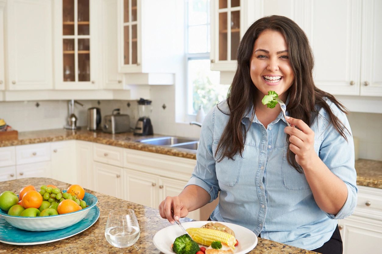 dark haired lady eating a stem of broccoli in a neutral contemporary modern kitchen at an island.